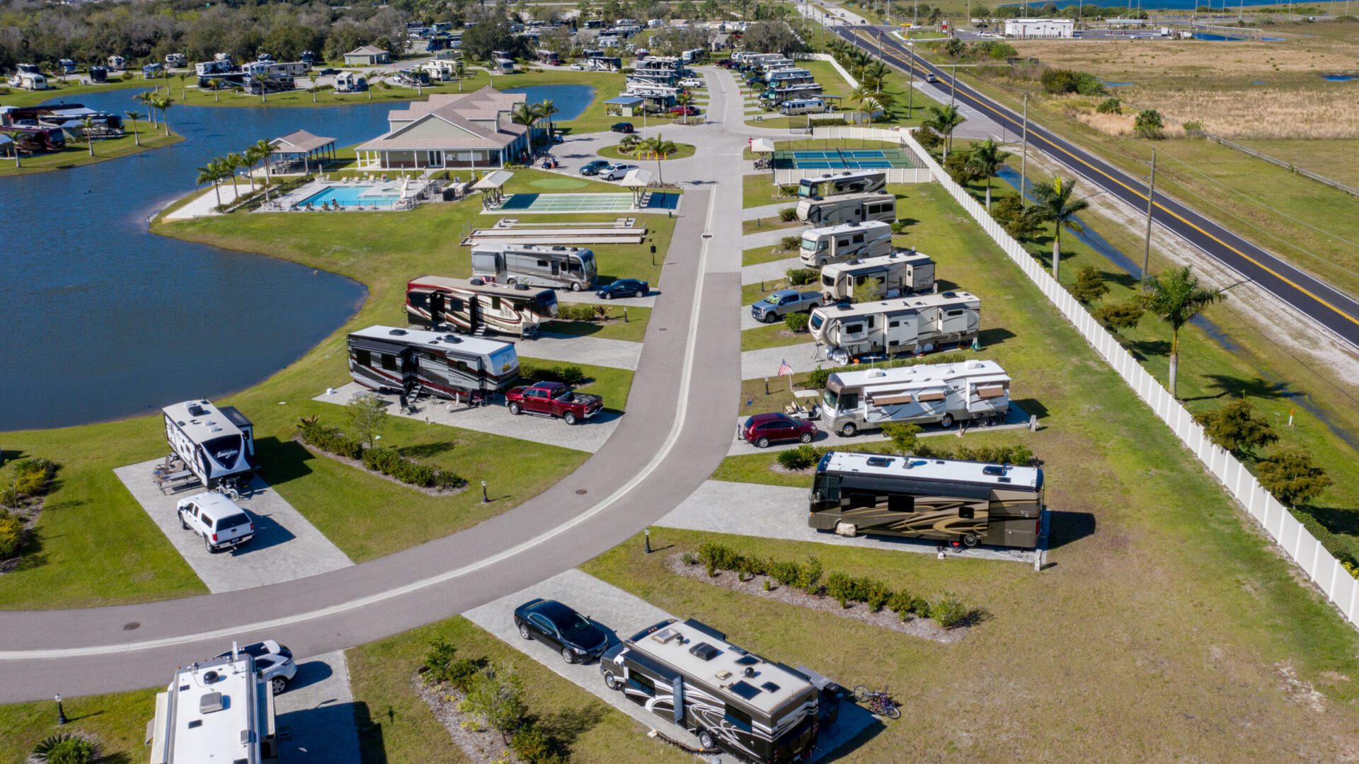 Aerial view of an RV park with several motorhomes parked on individual lots, a swimming pool, a lake, and a road running alongside the park.