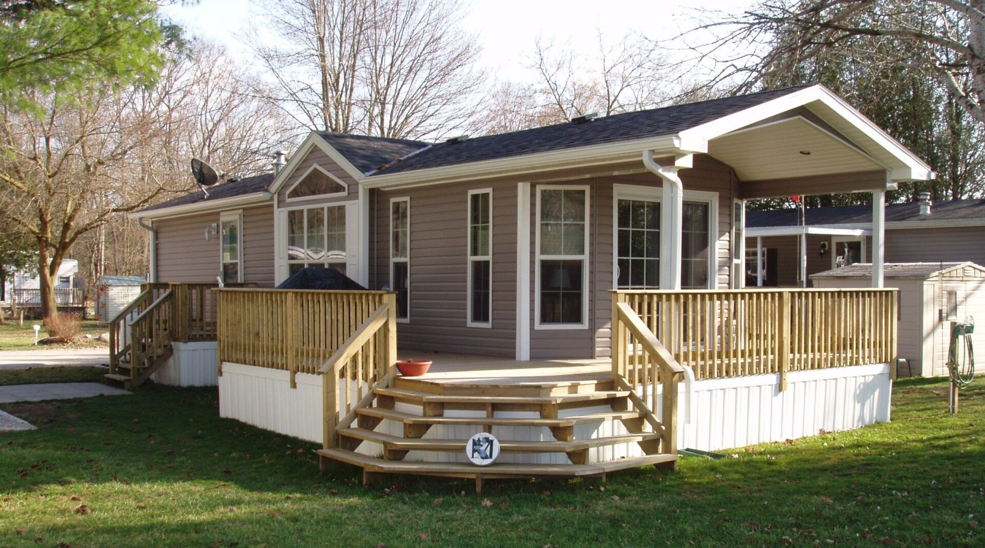 Single-story manufactured home with beige siding, white trim, and a covered front porch with wooden railings and steps, set on a green lawn with trees in the background.