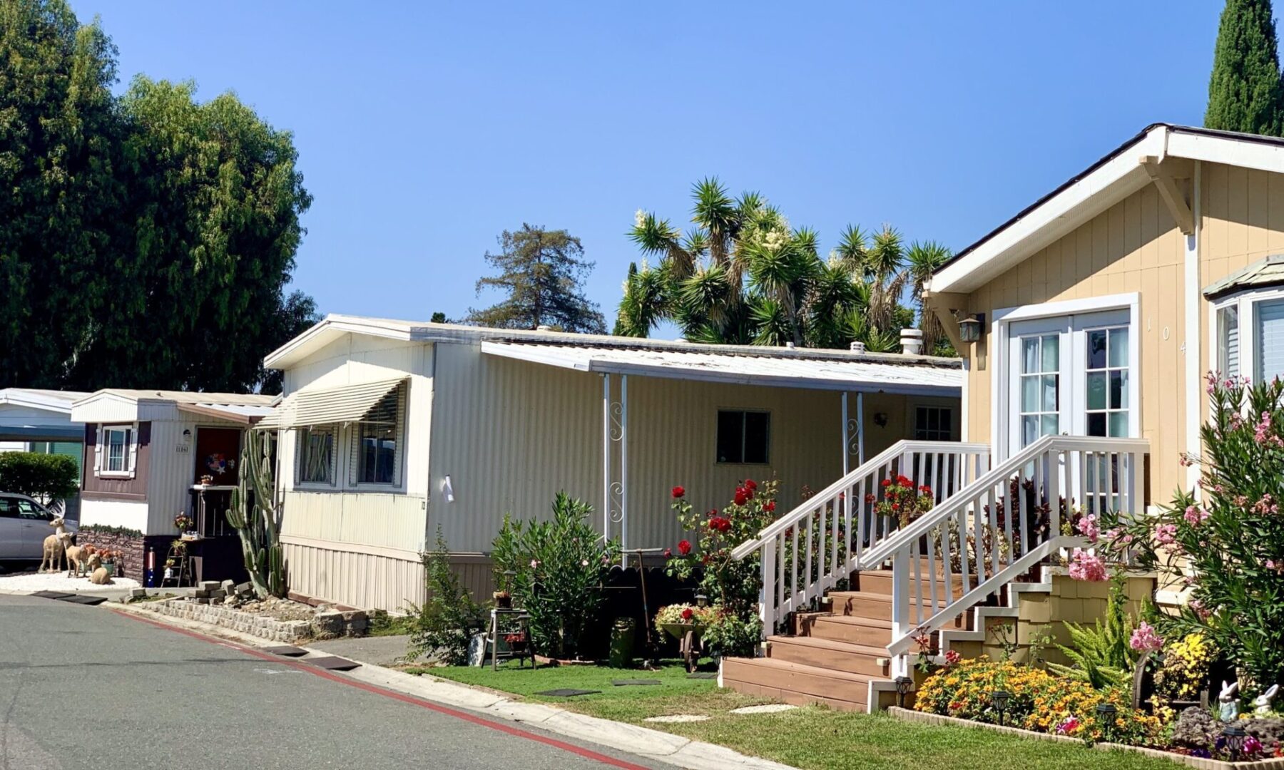 A row of manufactured homes with small gardens and flowers along a paved street on a clear, sunny day.