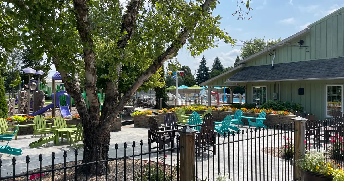 Patio area with colorful chairs, a tree, and flowers near a fenced pool, playground, and green building on a sunny day.