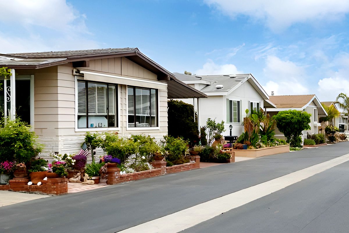 Row of single-story manufactured homes with well-kept gardens along a paved street under a partly cloudy sky.