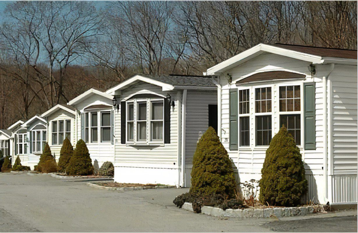 Row of white mobile homes with small bushes in front, situated along a paved road with leafless trees in the background.