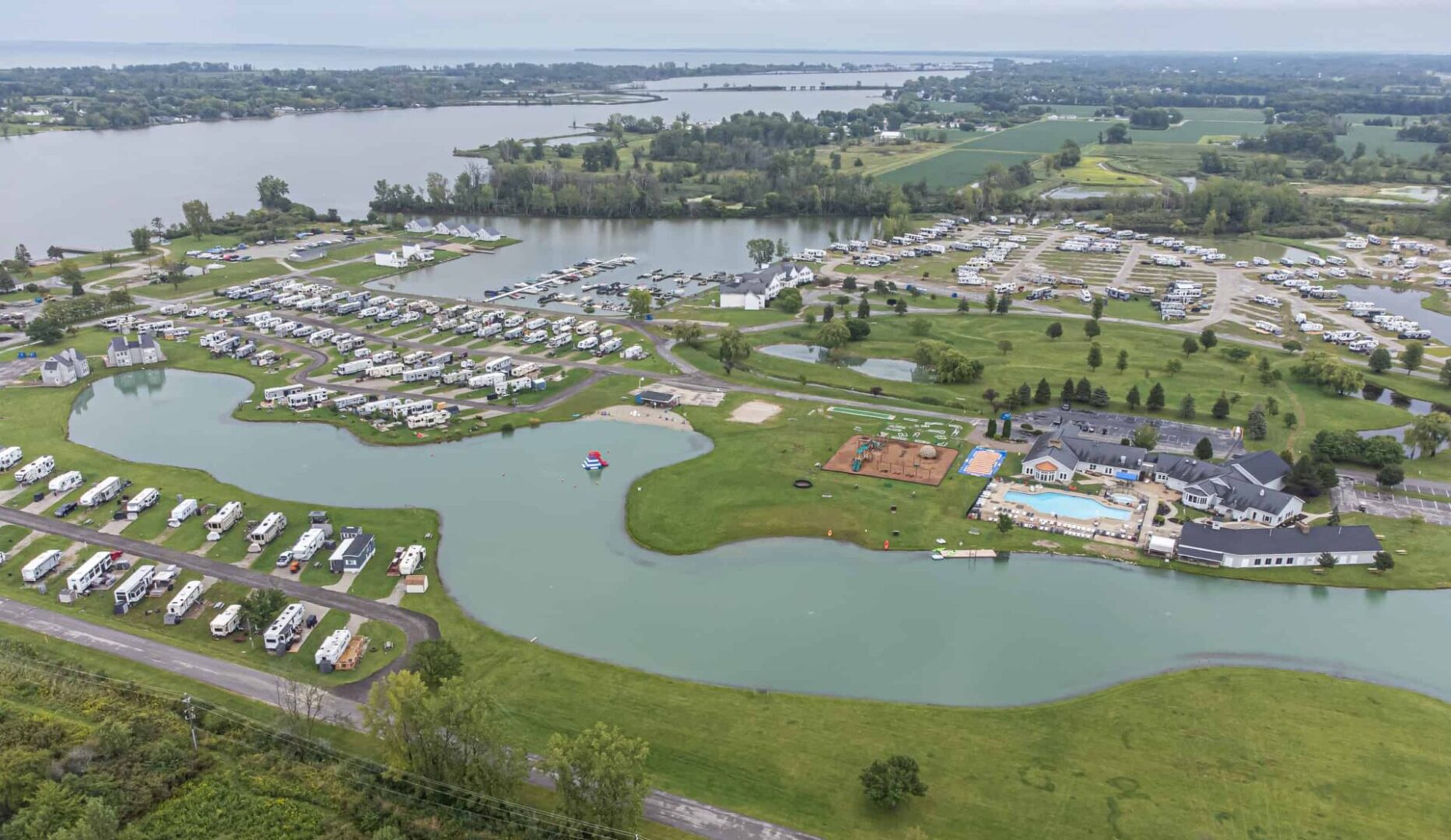 Aerial view of a large campground with rows of RVs, a swimming pool, ponds, and recreational areas near a lake, surrounded by green fields and trees.