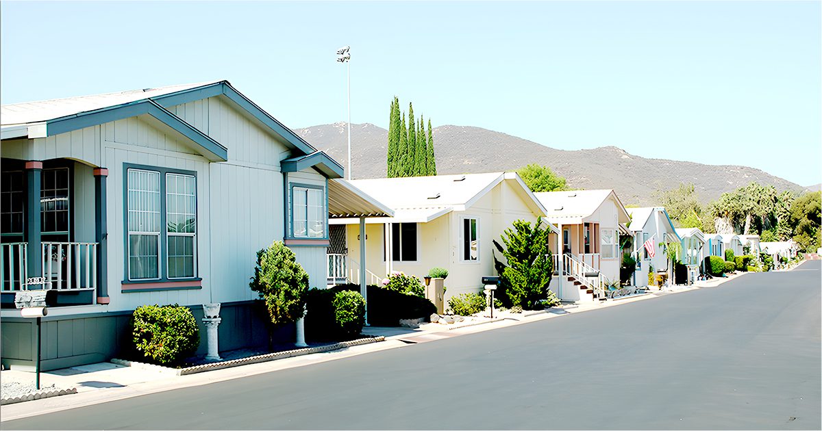 Row of manufactured homes with small front porches, neatly landscaped yards, and a paved street, set against a backdrop of hills and clear sky.