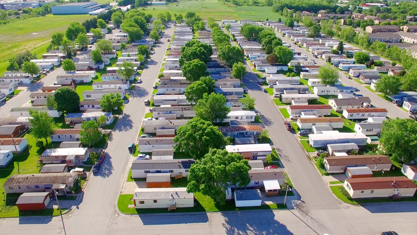 Aerial view of a mobile home park with rows of manufactured homes, streets lined with green trees, and open grassy areas in the background.