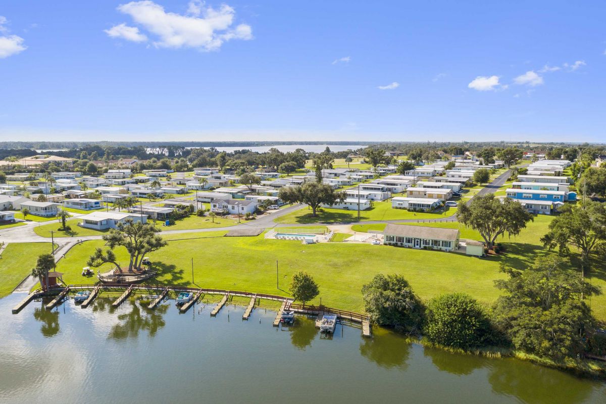 Aerial view of a lakeside mobile home park with green lawns, boat docks, and several rows of white homes under a clear blue sky.