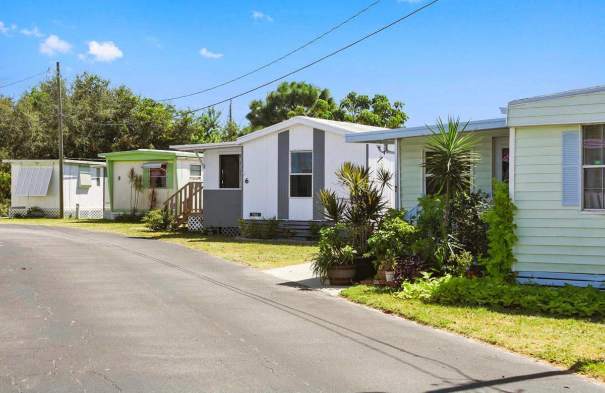 A row of small, single-story mobile homes with plants in front, situated along a paved road under a clear blue sky.