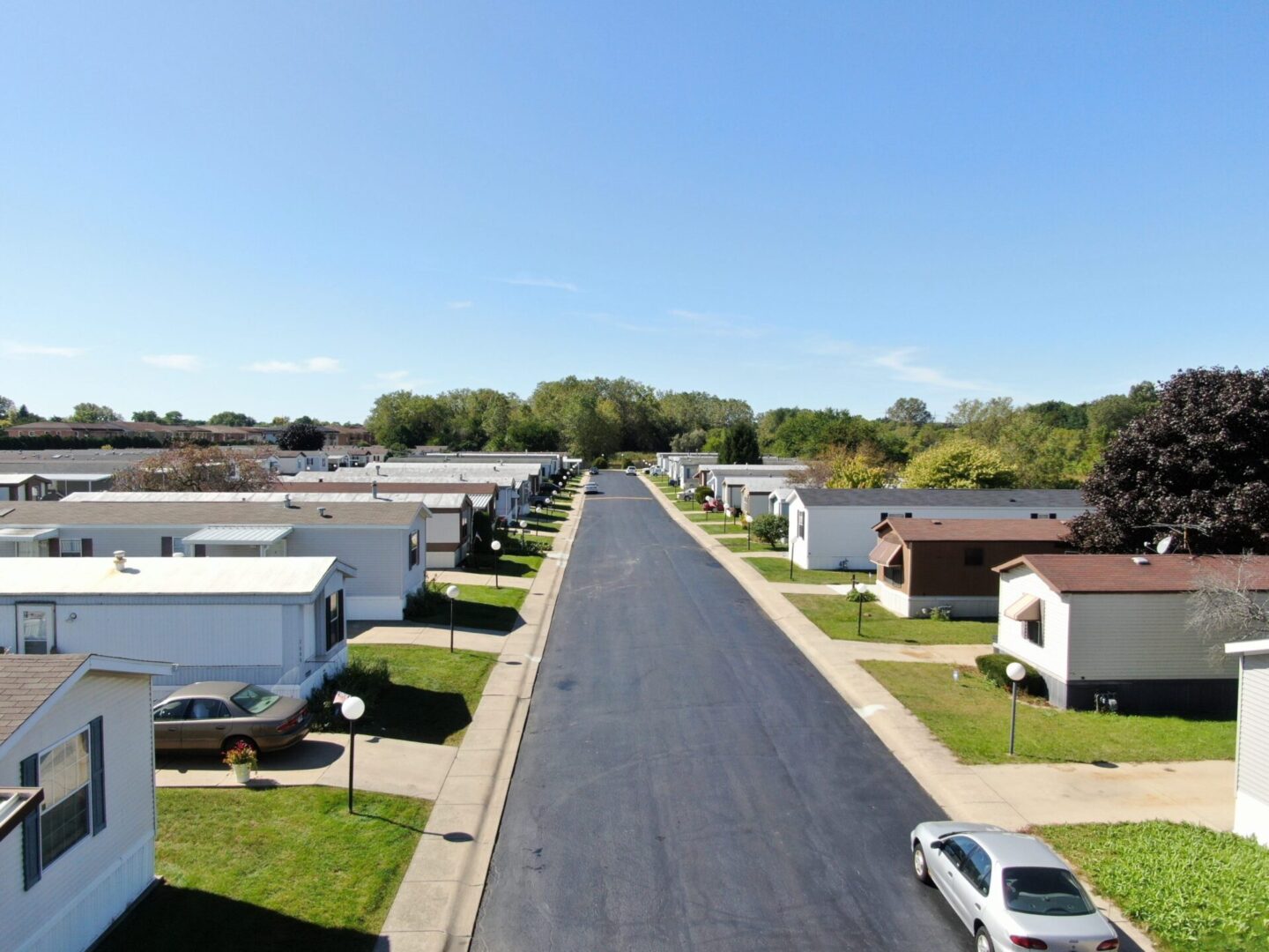 A paved street runs between rows of white and beige mobile homes with driveways, parked cars, and well-kept lawns under a clear blue sky.