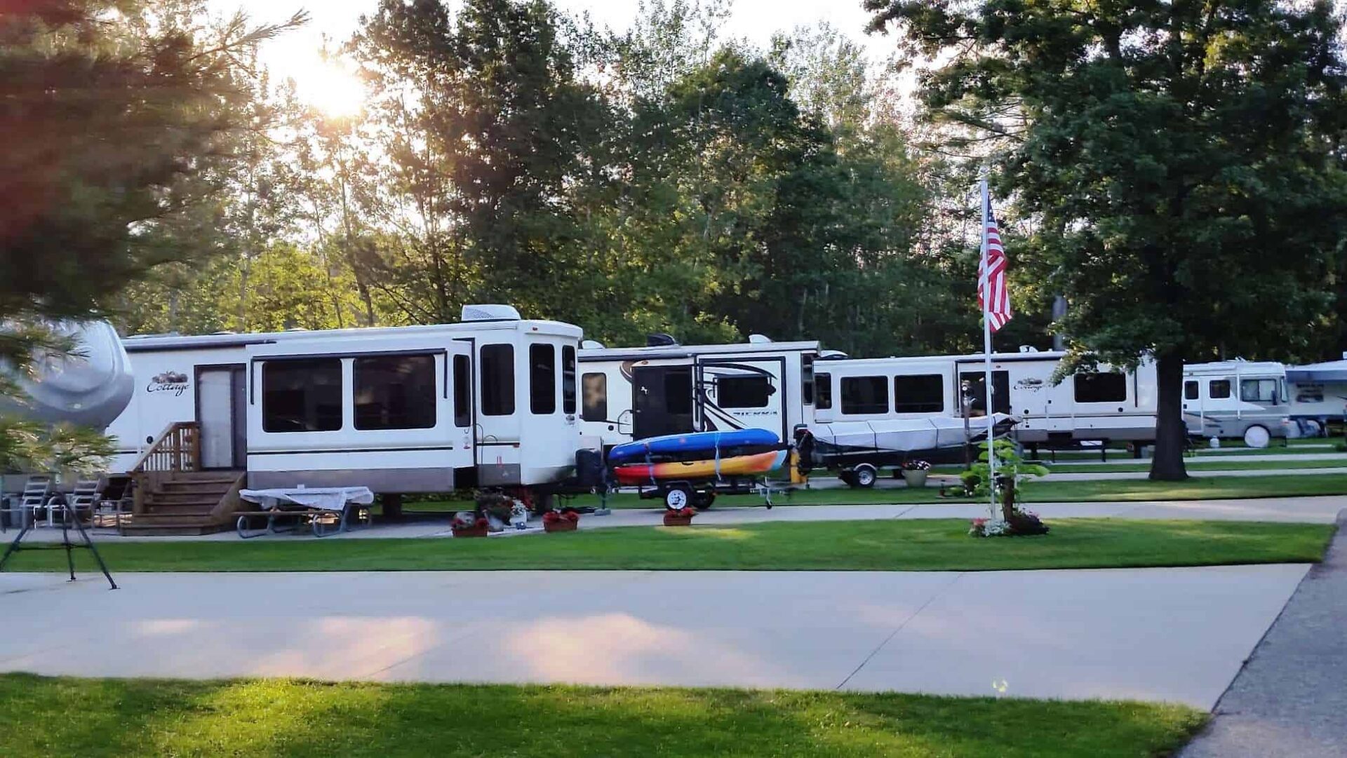 Several recreational vehicles are parked on a grassy lot with paved driveways, surrounded by trees. An American flag is displayed near the center.