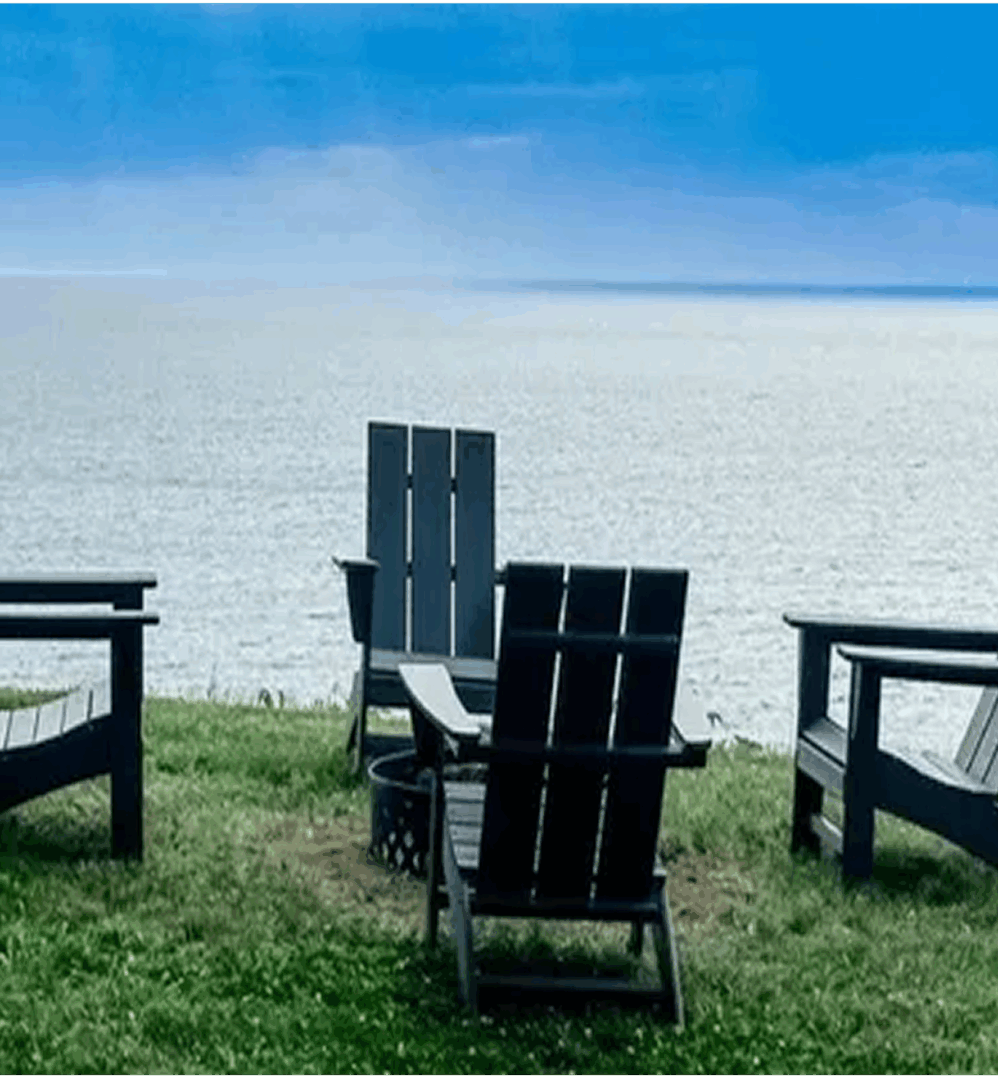 Empty black Adirondack chairs overlooking a calm lake under a blue sky.
